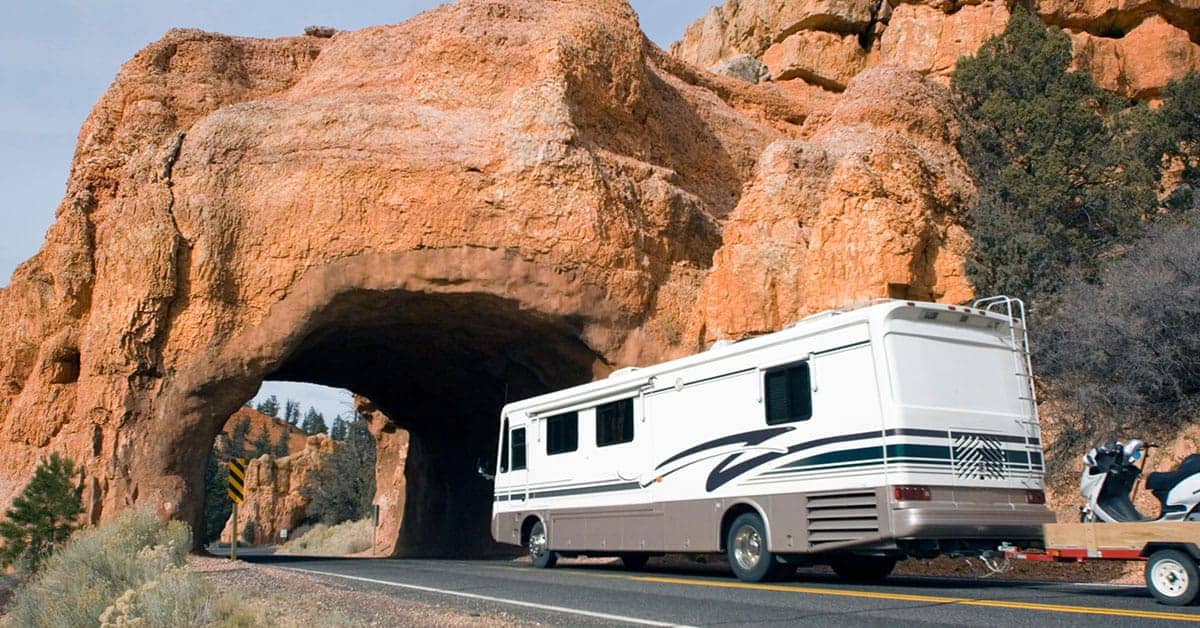 RV driving through a natural rock arch in a scenic landscape, emphasizing RV travel and maintenance importance.