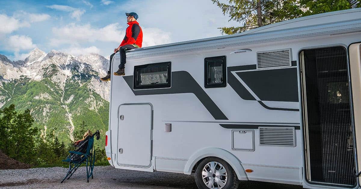 Man sitting on the roof of a modern RV in a scenic mountain landscape, illustrating outdoor RV lifestyle and maintenance awareness.