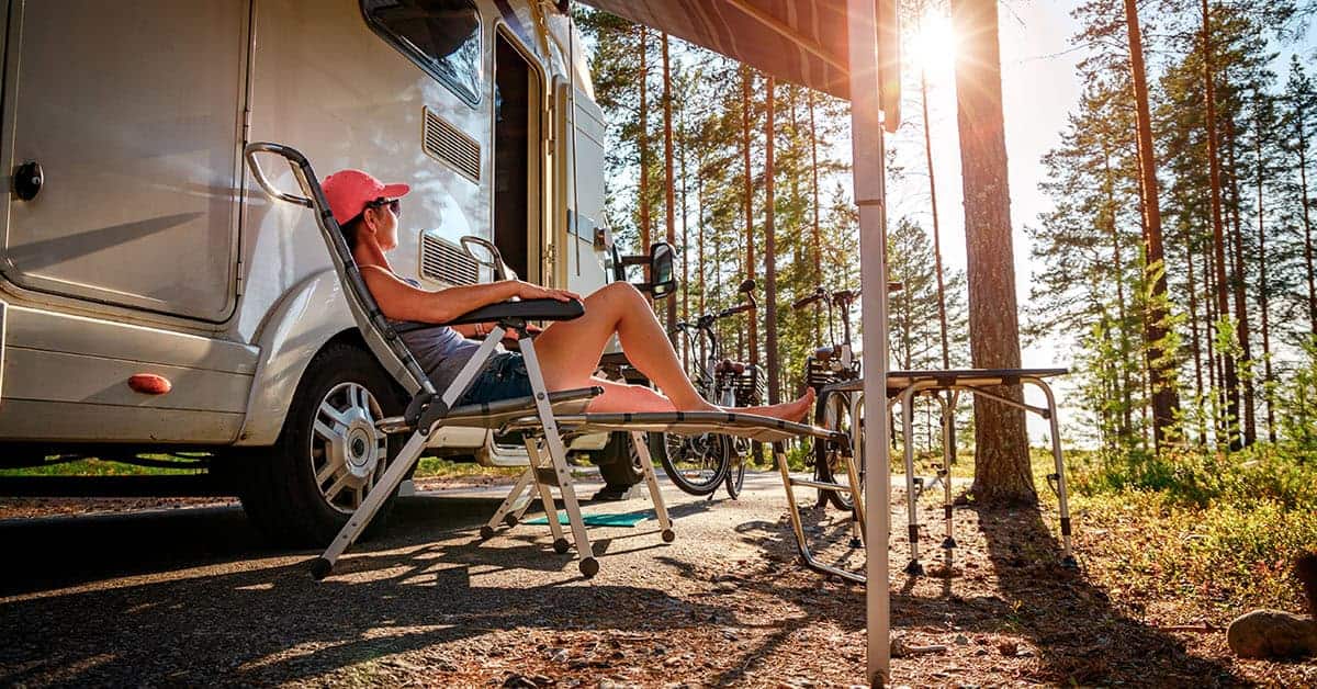 Woman relaxing in a camping chair beside a parked RV in a forested area, emphasizing RV lifestyle and leisure.