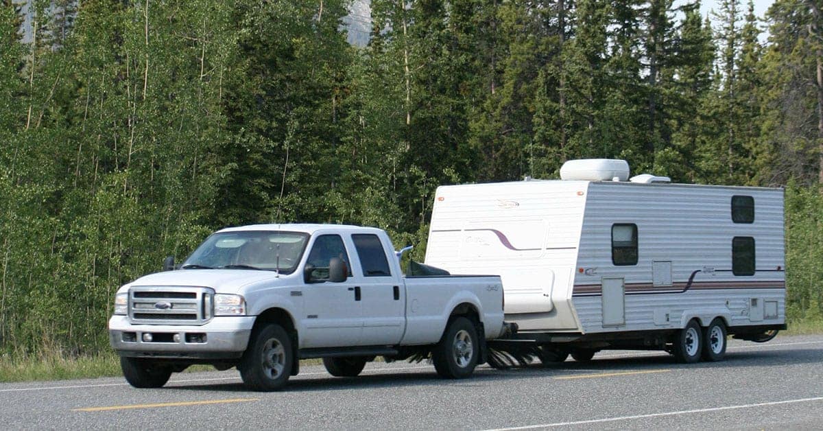 White pickup truck towing a travel trailer on a rural road surrounded by trees, representing trailer maintenance and travel.