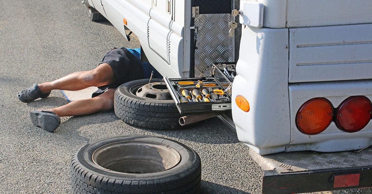 Person performing RV maintenance under vehicle with tools and spare tire visible.